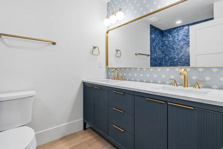 Dual sink vanity with gold-tone hardware, rectangular mirror with gold-tone frame, two-tone patterned wall tile, and light wood-finish flooring