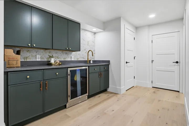 Hardwood flooring with a wet bar featuring dark green cabinetry, gold hardware, a built-in beverage refrigerator, and a gold-tone faucet with a patterned backsplash