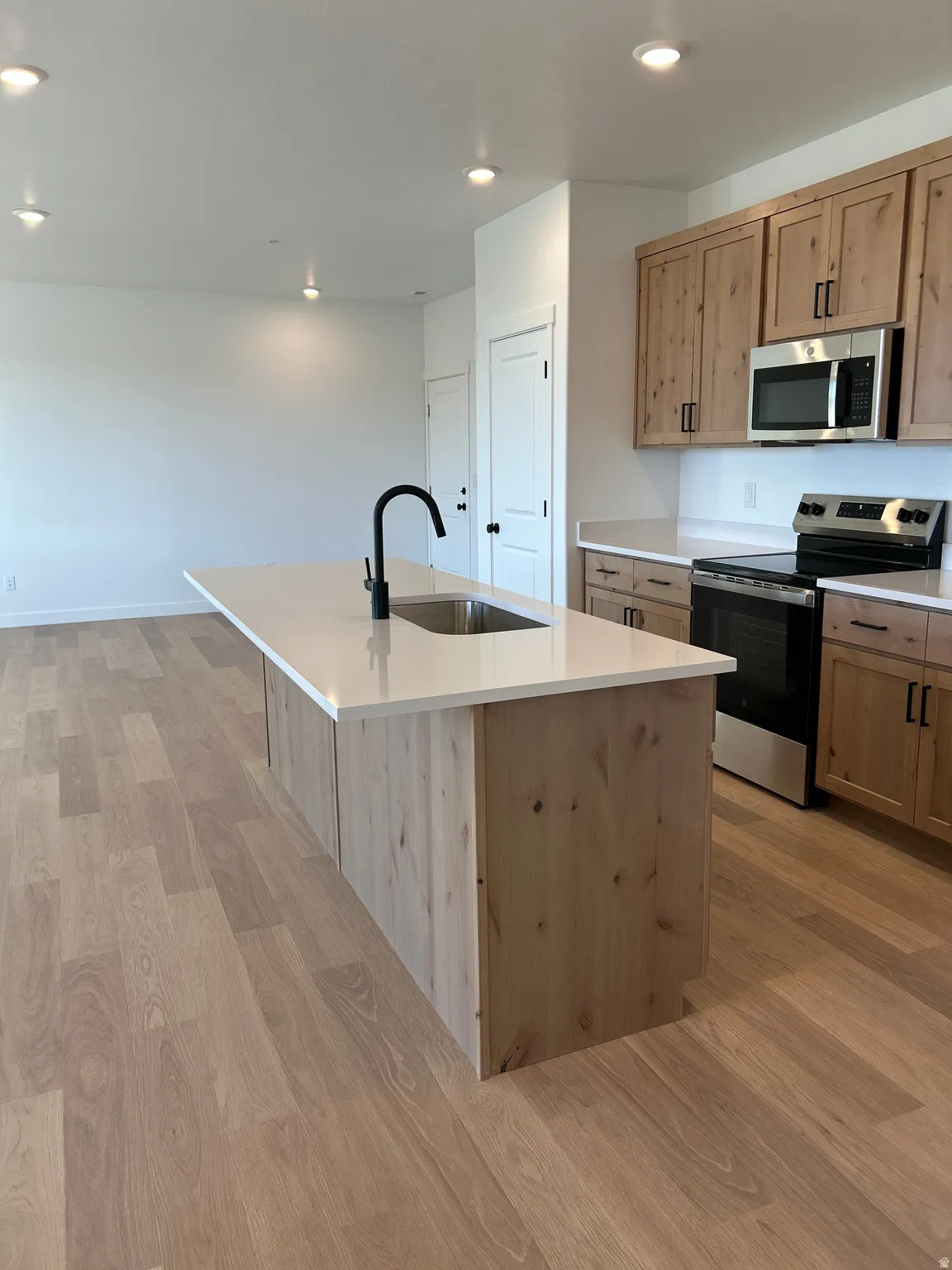 Kitchen featuring wood-finish flooring, an island with an integrated sink, a matte black faucet, light wood cabinetry, and stainless steel appliances