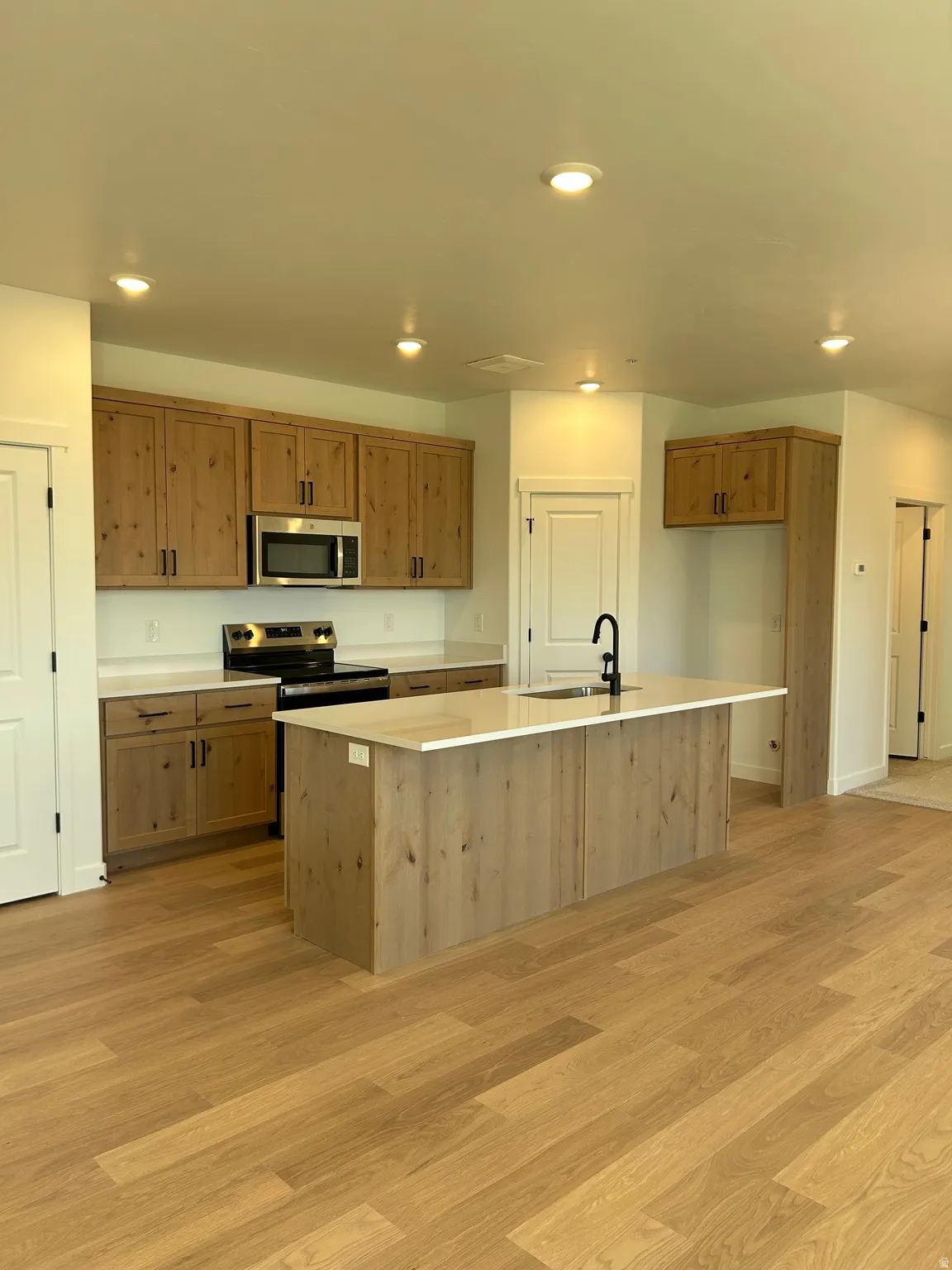 Kitchen featuring wood-finish cabinetry, stainless steel appliances, a center island with sink, light-toned countertops, and recessed lighting