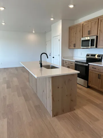 Kitchen featuring wood-finish flooring, an island with an integrated sink, a matte black faucet, light wood cabinetry, and stainless steel appliances