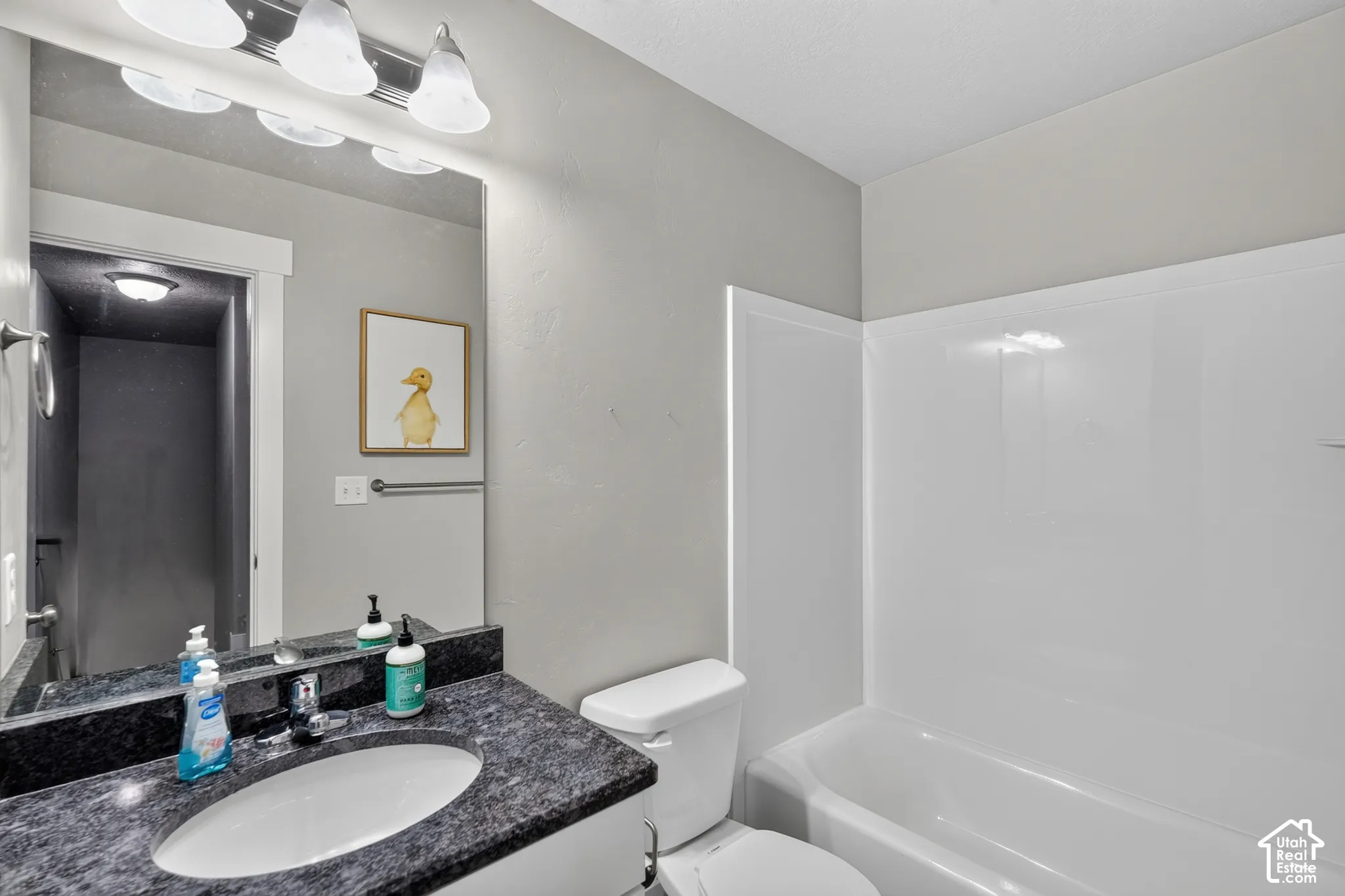 Bathroom featuring a vanity with a stone-finish countertop, undermount oval sink, chrome faucet, large vanity mirror with a multi-bulb light fixture, and a white tub-shower surround