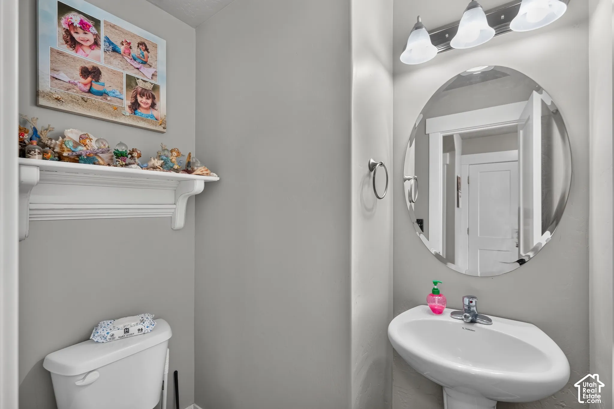 Pedestal sink, wall-mounted oval mirror, three-light vanity fixture, and a white wall shelf