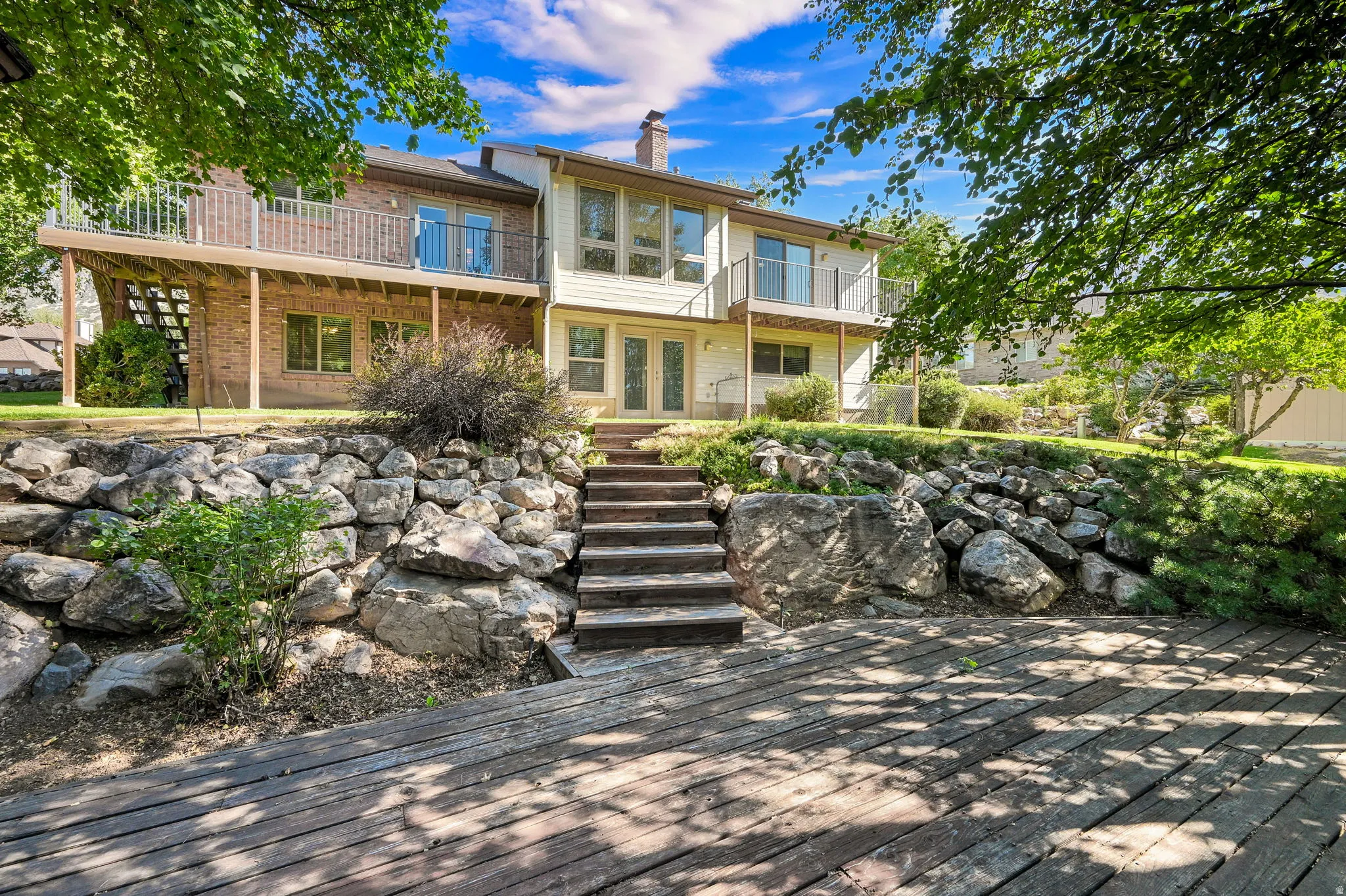 Expansive wooden deck, brick and siding exterior of home, rock retaining wall, exterior stairs, and ground-level patio doors.