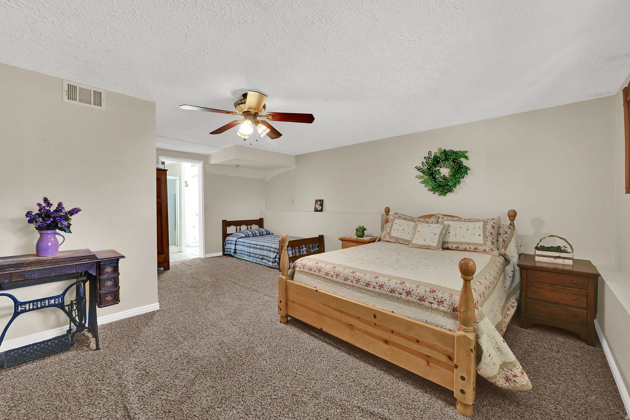 Large bedroom with carpeting, light neutral wall paint, and a ceiling fan with lighting. Bedroom has it's own private bathroom