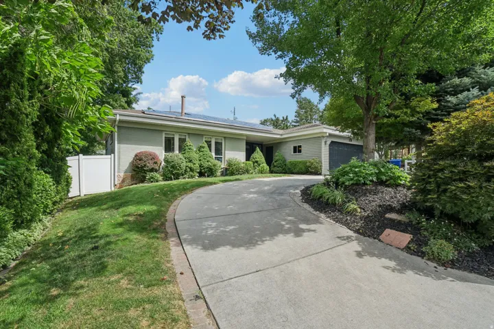Curved concrete driveway, established landscaping with mature trees, attached garage, and a white privacy fence