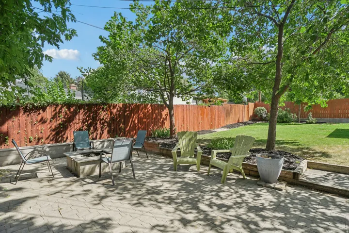 Paver patio with a built-in fire pit, surrounded by raised garden beds, a wood privacy fence, and mature trees