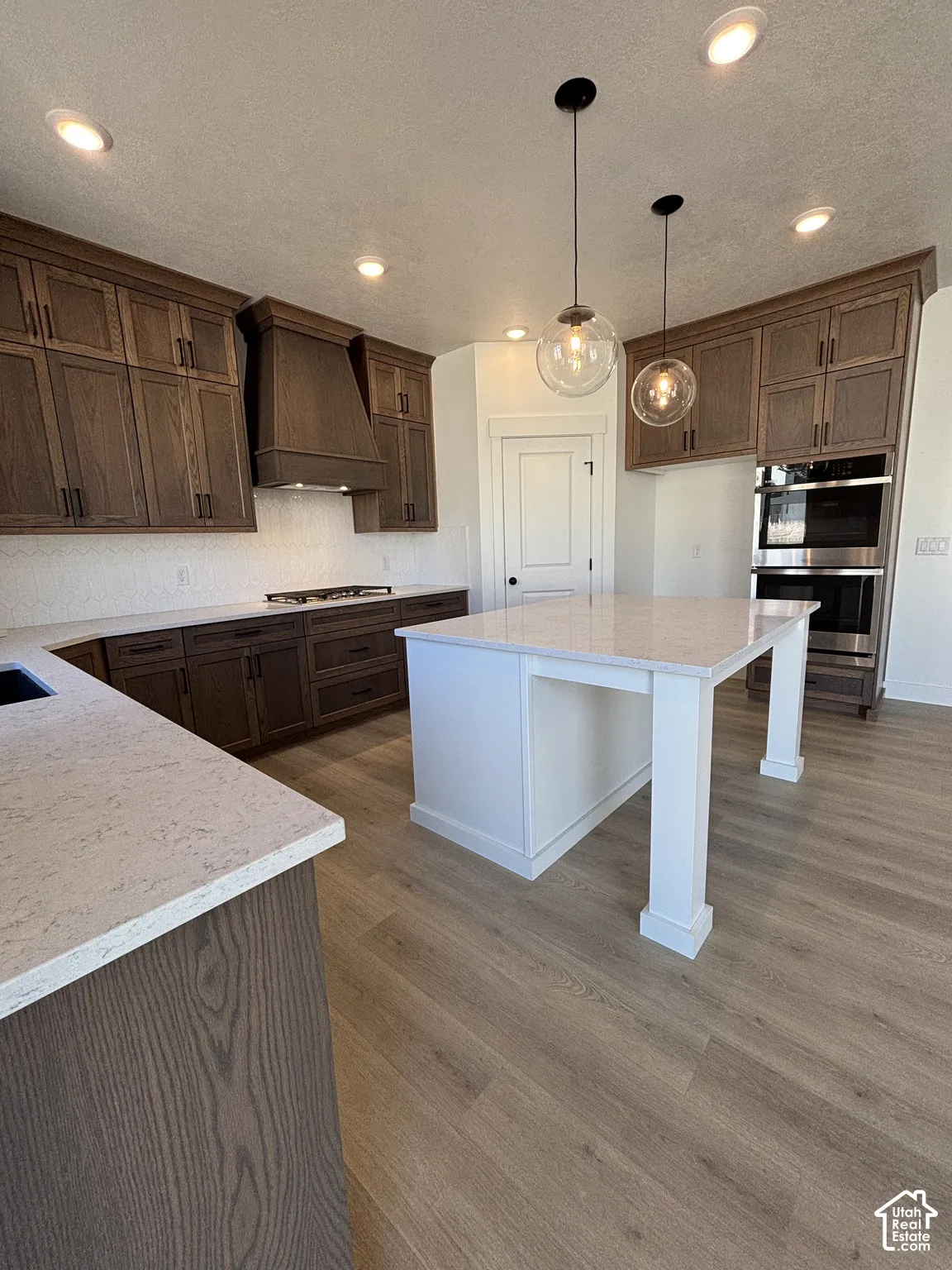 Modern kitchen featuring wood-finish cabinetry, light-toned countertops, a central island with pendant lighting, a built-in oven, and wood-finish flooring