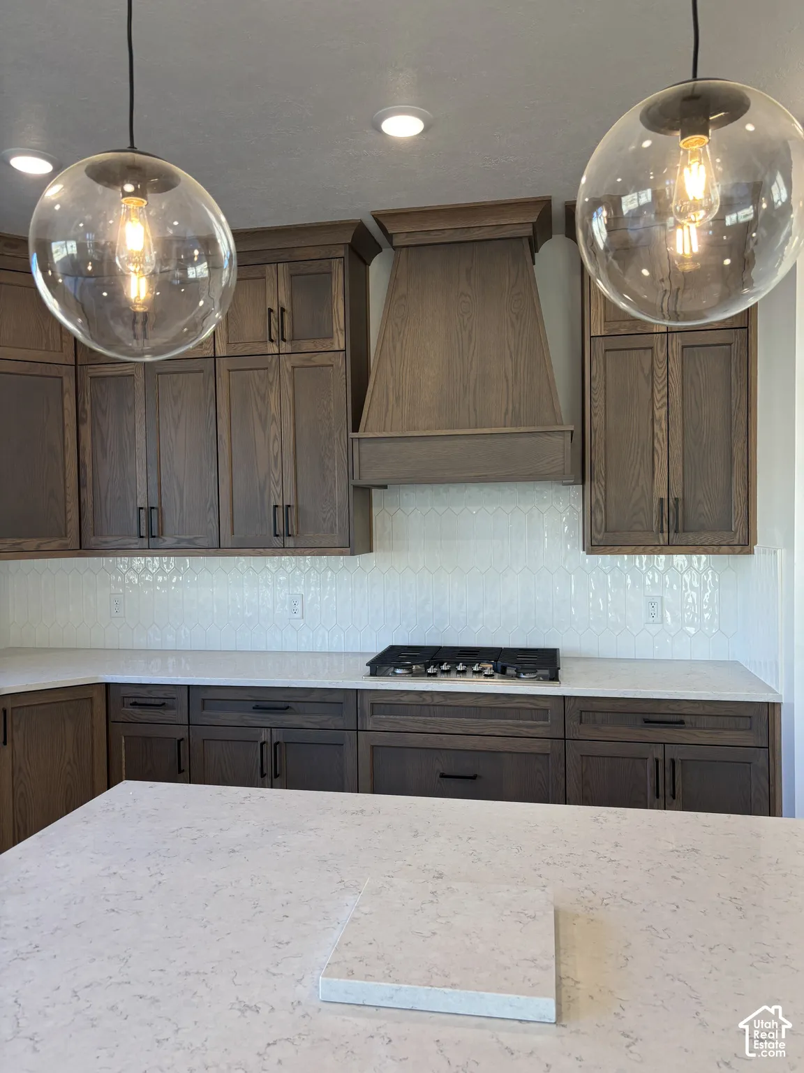 Recessed lighting and clear globe pendant lights illuminate the kitchen featuring dark wood cabinetry, a decorative wood hood, a gas cooktop, and light-toned countertops