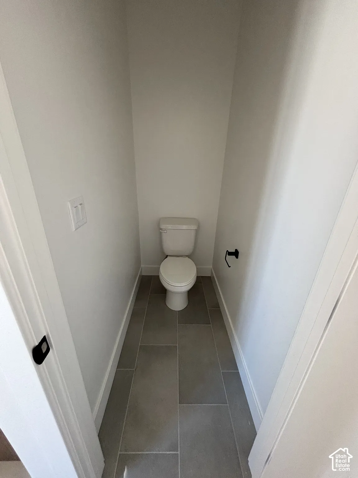 White porcelain toilet with a dark-finish toilet paper holder, neutral-toned wall paint, and gray rectangular floor tiles