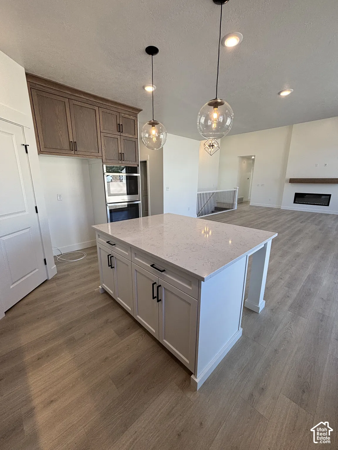 Kitchen island with a light-toned countertop, wood-finish flooring, recessed lighting, double wall oven, and pendant light fixtures