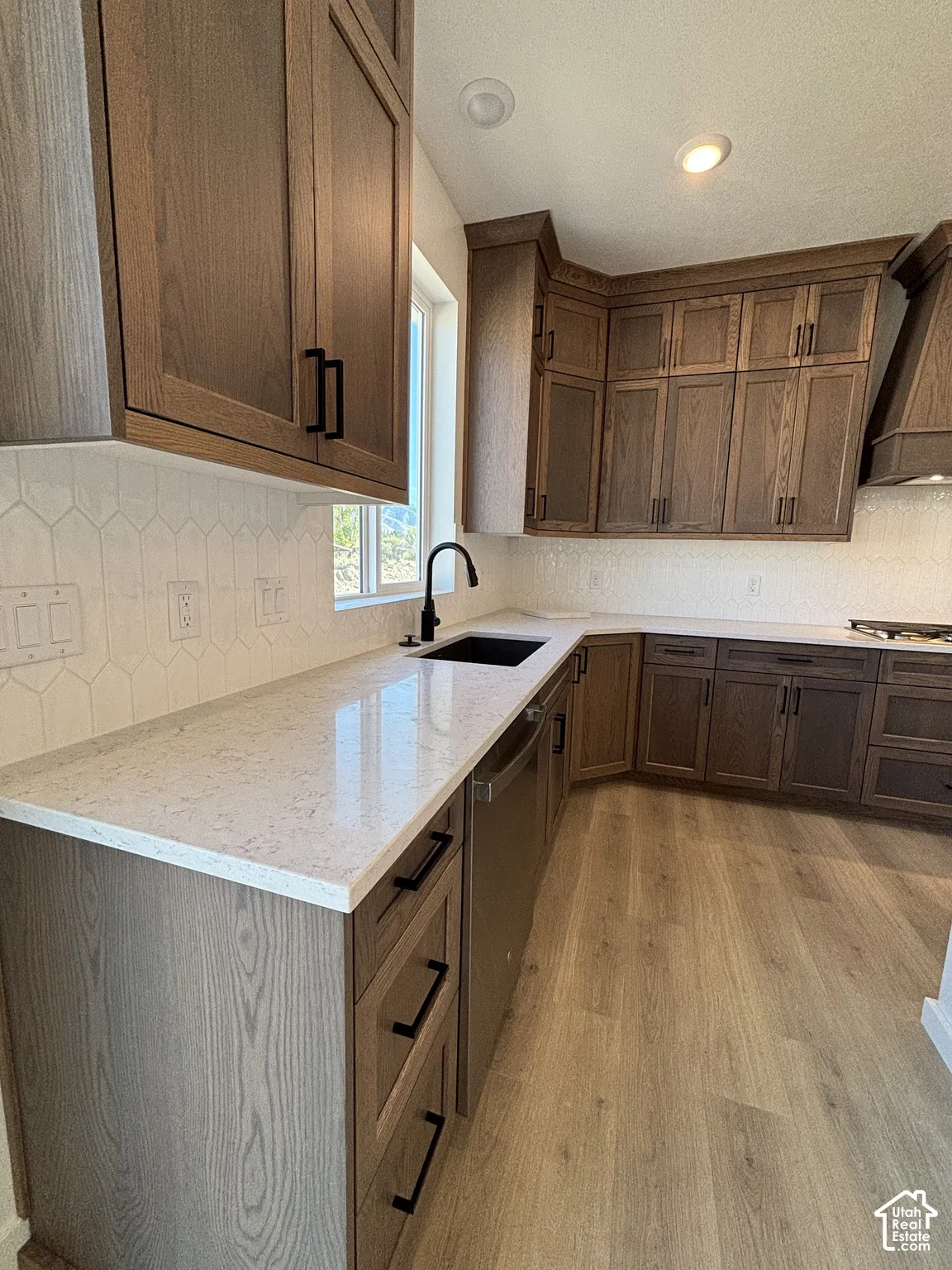 L-shaped kitchen featuring wood-finish cabinetry, light-toned countertops, a black sink with a gooseneck faucet, a stainless steel dishwasher, and wood-finish flooring