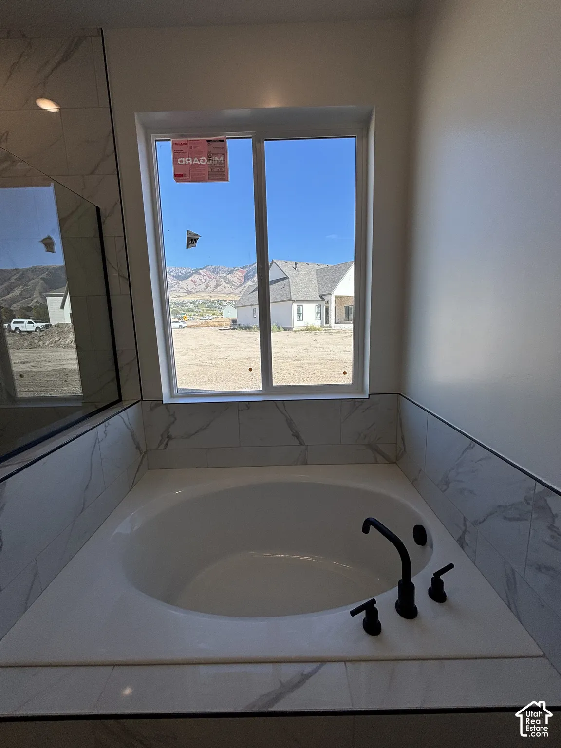Built-in soaking tub with matte black fixtures, surrounded by large format tile with a distinct veining pattern, and a large window