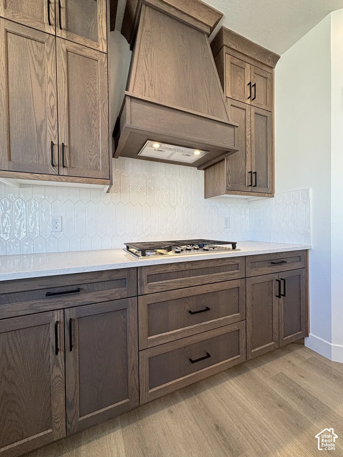 Wood-finish cabinetry with black hardware, white solid surface countertops, patterned tile backsplash, and wood-finish flooring