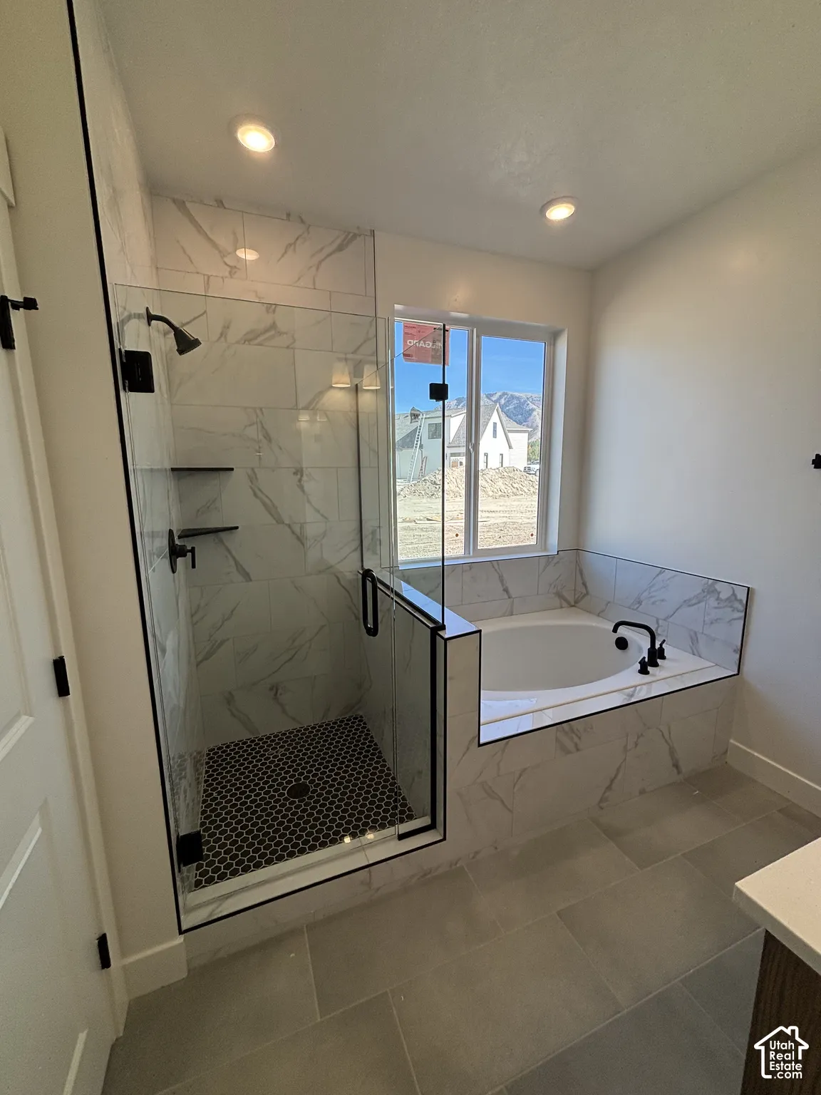 Spacious bathroom featuring a built-in soaking tub, glass-enclosed shower with black hexagonal tile, and large format floor tiling