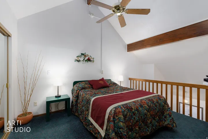 Vaulted white ceiling, wood-finish ceiling fan, exposed wood-tone beam, wood-tone railing, and blue carpeting