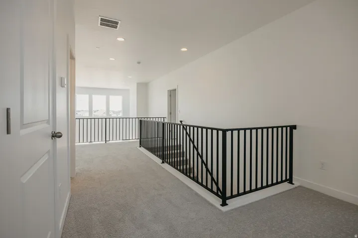 Carpeted hallway with white walls, recessed lighting, and a black metal staircase railing