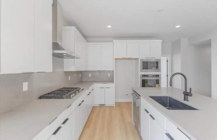 Modern kitchen featuring light wood-finish flooring, white flat-panel cabinetry, a stainless steel gas cooktop with coordinating hood, and light-toned countertops
