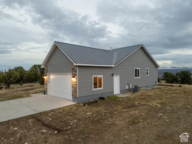 Gray siding exterior with a metal roof, stone accents, attached garage with concrete driveway, and wall-mounted exterior lighting with side door to the garage.