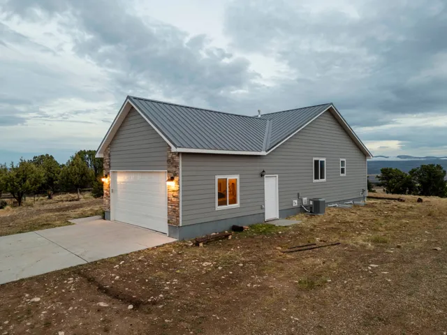 Gray siding exterior with a metal roof, stone accents, attached garage with concrete driveway, and wall-mounted exterior lighting with side door to the garage.