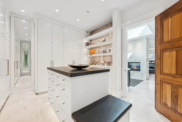Custom closet featuring white cabinetry, a central island with a dark countertop, integrated shelving with accent lighting, and light-toned tile flooring