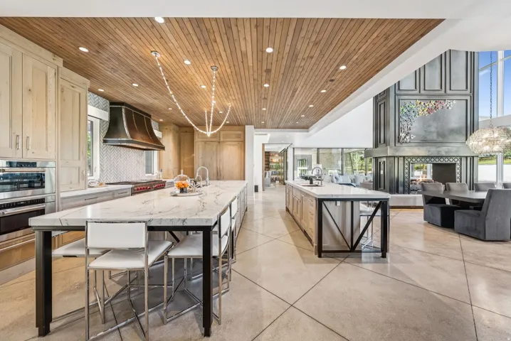Gourmet kitchen and living area featuring a wood-panel ceiling with recessed lighting, two large islands with stone countertops, integrated stainless steel appliances, and a substantial fireplace with a decorative mantel