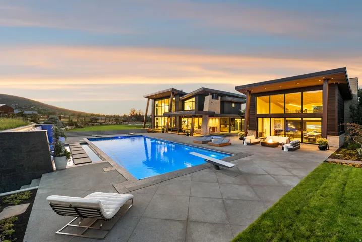 Modern architectural residence featuring a large rectangular swimming pool with a diving board, expansive patio space, integrated outdoor lighting, and a manicured lawn