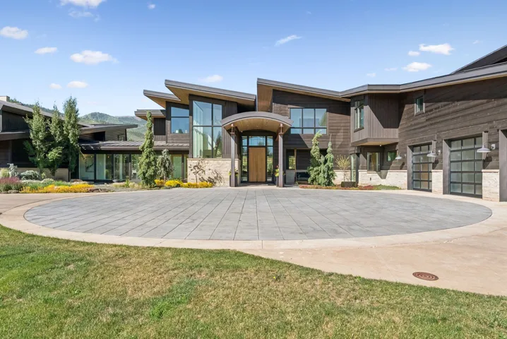 Contemporary home with a circular paver driveway, dark wood siding, stone accents, multiple large windows, and integrated garage doors