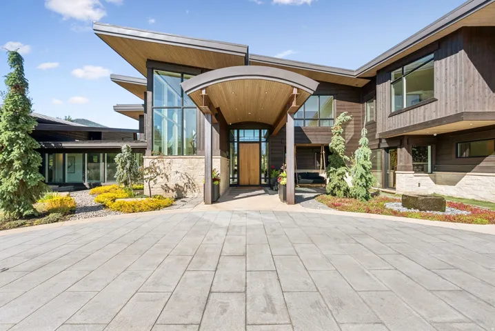 Contemporary architectural residence featuring a wood-clad exterior, extensive glass panels, a curved portico entry, a stone-faced foundation, and a paver driveway
