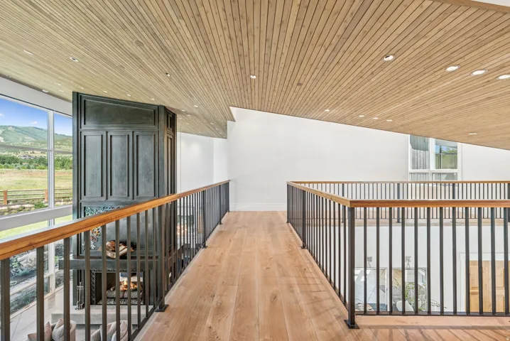 Elegant upper-level hallway featuring warm wood-finish flooring, a vaulted wood-slat ceiling with recessed lighting, and dual metal railings with wood top rails