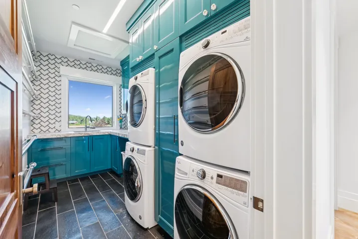 Laundry room featuring stacked washer and dryer units, a utility sink with a gooseneck faucet, teal cabinetry, and dark rectangular floor tiling