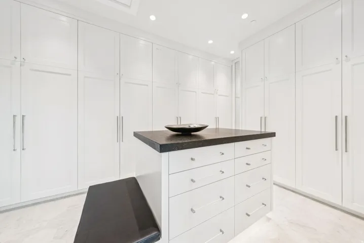 Luxurious walk-in closet featuring floor-to-ceiling white cabinetry with chrome hardware, a central island with dark wood-finish countertop and integrated vessel sink, recessed ceiling lighting, and light wood-finish flooring throughout