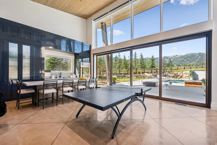 Open-concept recreation space showcasing soaring wood-ceiling architecture, floor-to-ceiling multi-panel glass walls framing mountain and vineyard vistas, polished concrete flooring, a built-in wet bar with cabinetry and countertop workspace, and a regula
