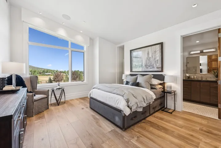 Bright bedroom featuring a large picture window with a scenic outdoor landscape, light wood-finish flooring, recessed lighting, and a private en-suite bathroom with a dark wood vanity
