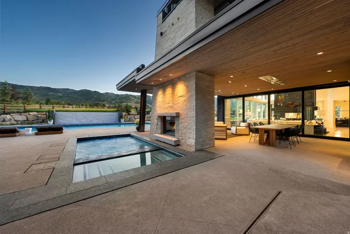 Modern luxury outdoor living space featuring a resort-style heated spa and swimming pool with integrated lighting, expansive paved terrace in neutral stone, a substantial covered pavilion with fireplace and ambient recessed lighting, floor-to-ceiling glas