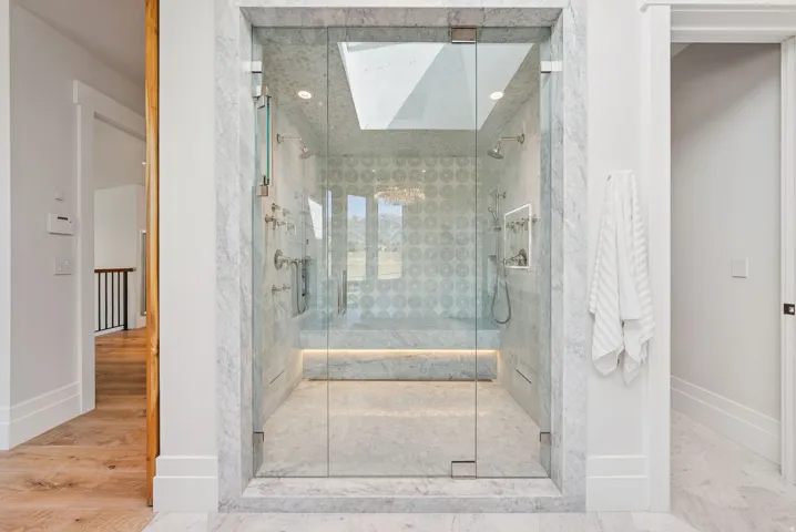 Luxurious spa-inspired shower enclosure featuring frameless glass doors, marble-look tile surround with geometric patterning, a spacious walk-in layout with integrated bench seating, polished chrome fixtures, and a skylight providing natural illumination