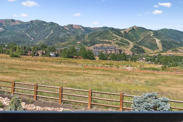 Expansive mountain landscape featuring a split-rail fence, open fields, distant buildings, and a clear sky