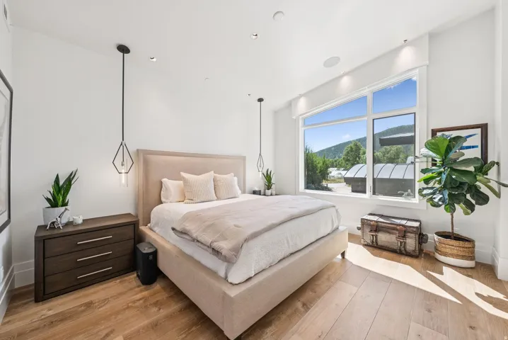 Bright, light-filled bedroom featuring a wood-finish upholstered platform bed with coordinating nightstands, pendant lighting, and expansive windows with mountain and landscape vistas