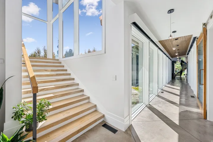 Modern staircase featuring alternating wood-finish and concrete treads with a sleek metal handrail, illuminated by expansive multi-pane windows offering abundant natural light and skyline views