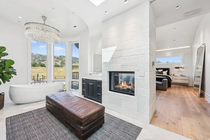 Luxurious primary bathroom featuring a freestanding soaking tub, double-sided linear fireplace with white brick surround, triple-pane windows framing mountain landscape, wood-finish flooring, and seamless passage to adjoining bedroom suite