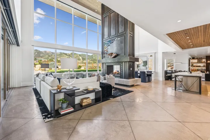Spacious living area featuring floor-to-ceiling windows, a grand fireplace with decorative paneling, wood-finish flooring, and high ceilings