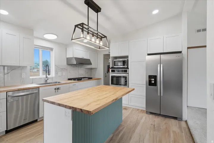 White shaker cabinetry, stainless steel appliances, wood-finish flooring, island with wood countertop, and modern pendant lighting