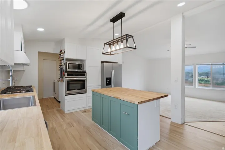 Modern kitchen featuring a wood-finish island with teal cabinetry and natural wood countertop, stainless steel appliances including dual wall ovens and refrigerator with ice and water dispensers, pendant lighting with metal fixtures, light wood-finish flo