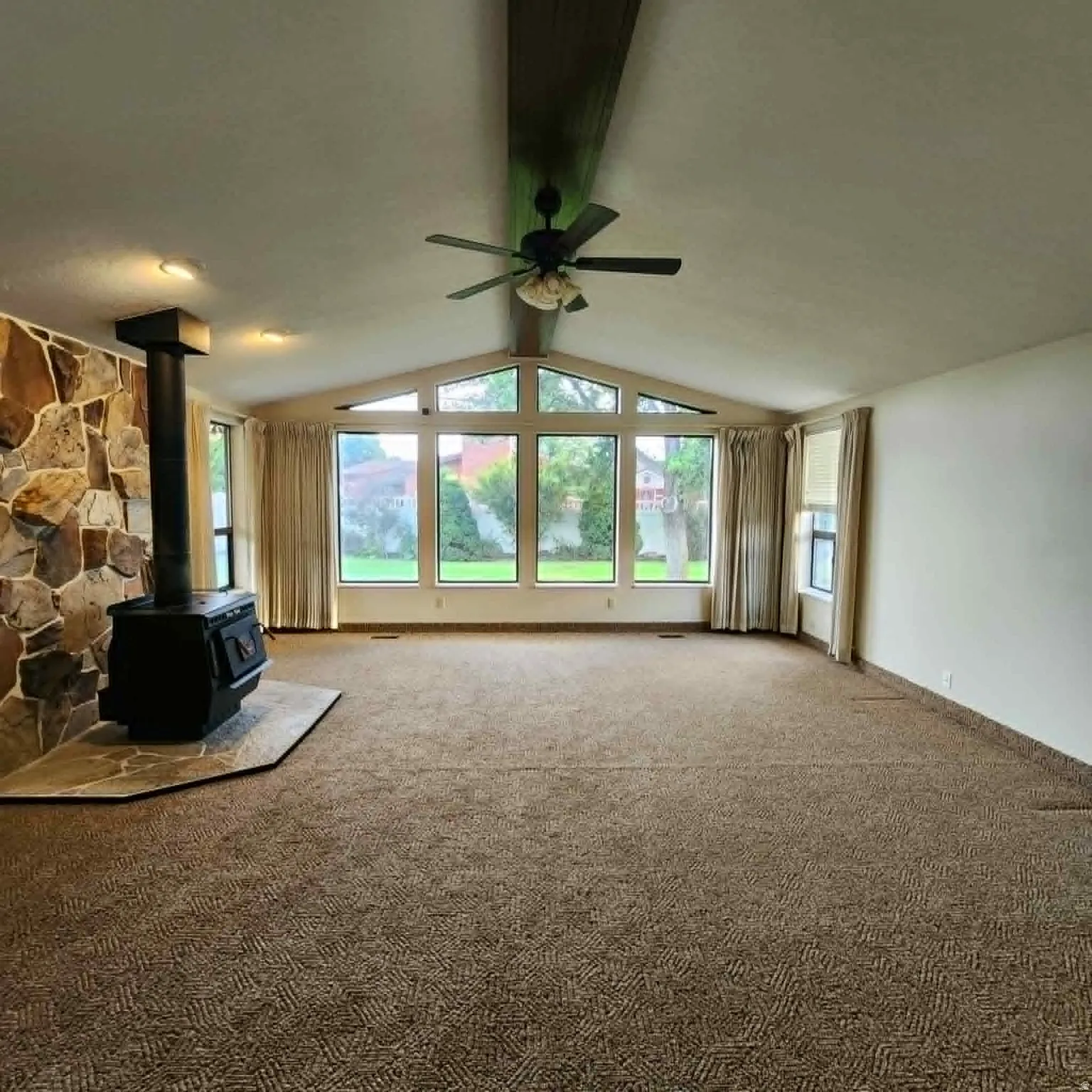 Vaulted ceiling with an exposed beam, stone-clad wood stove, large picture windows, ceiling fan, and patterned carpet flooring