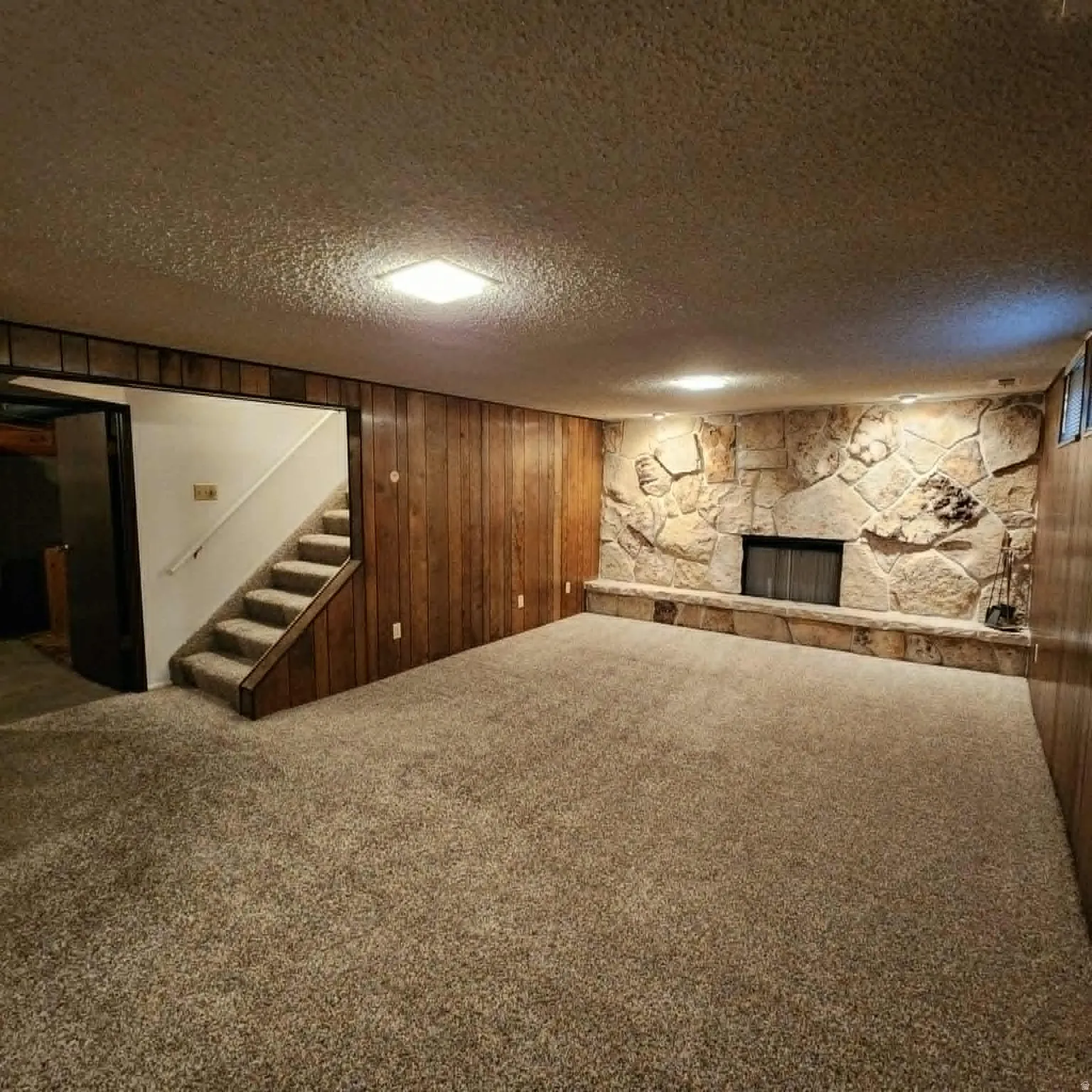 Expansive carpeted area featuring a stone fireplace with hearth, wood paneling, and an exposed staircase