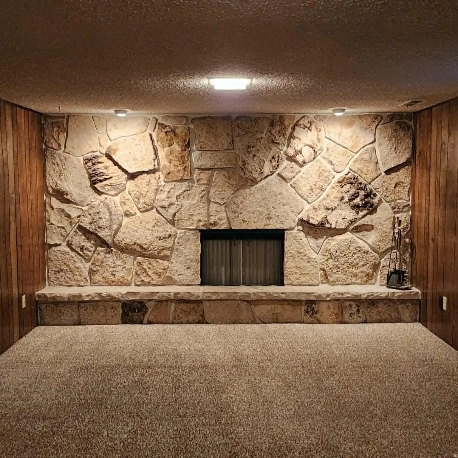 Stone fireplace with a hearth, integrated firebox, wall-mounted lighting, and carpeted flooring