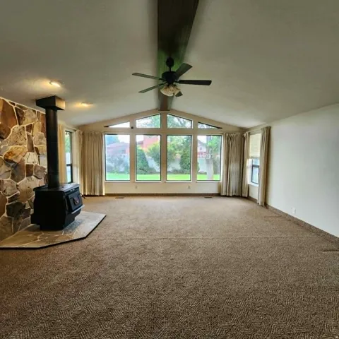 Vaulted ceiling with an exposed beam, stone-clad wood stove, large picture windows, ceiling fan, and patterned carpet flooring