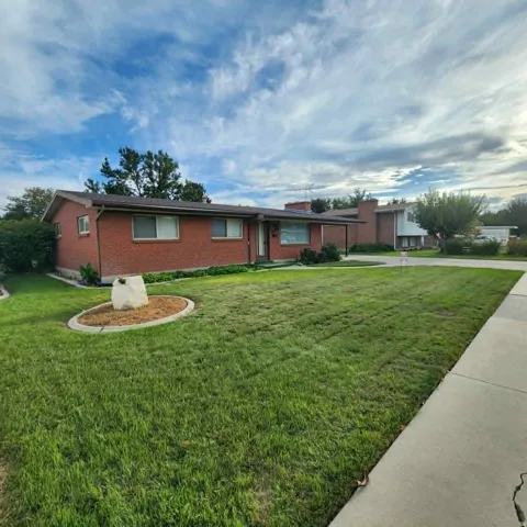 Red brick exterior with a dark roof, front yard with green grass, established landscaping, and a concrete sidewalk