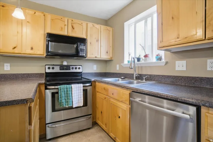 Light wood cabinetry, dark countertop surfaces, stainless steel appliances, and a double basin sink with a window above