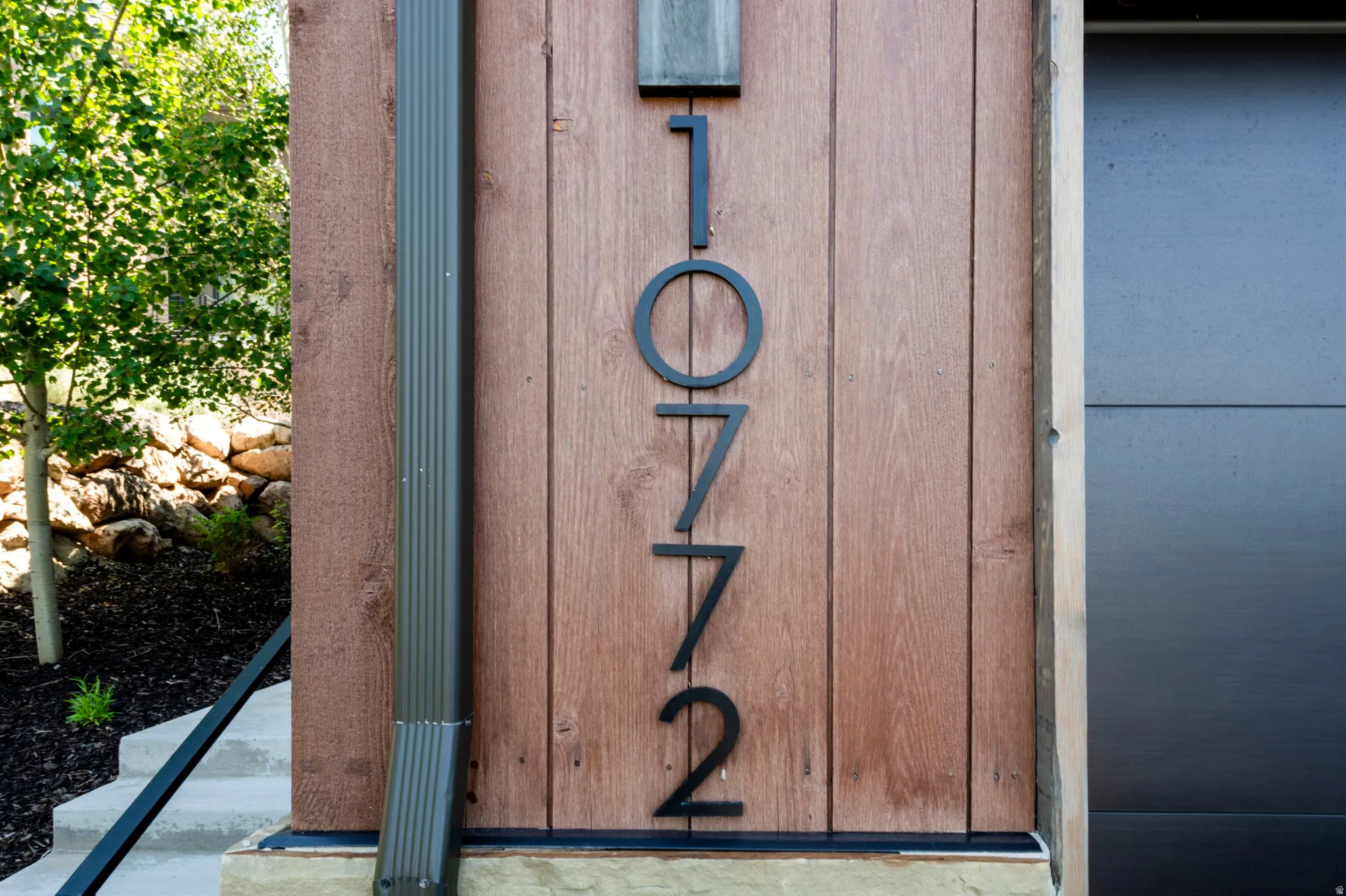 Vertical wood-finish siding with large black house numbers, dark gray downspout, and concrete steps with a black metal railing